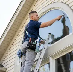 Professional window cleaner on a ladder using a squeegee to clean an exterior arched window, Nashua, NH.